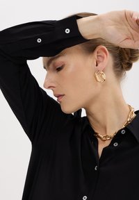 Camicia nera con bottoni argentati, abbinata a una collana di catena dorata spessa e orecchini a cerchio in oro testurizzati. L'illuminazione soffusa mette in evidenza i dettagli.