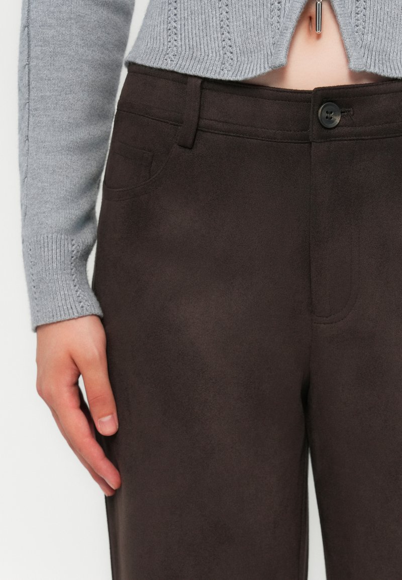 Zbliżenie ręki spoczywającej obok ciemno-brązowych spodni z zamszu z zapięciem na guziki, noszonych z jasnoszarym swetrem dzianym.