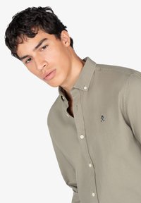 Camisa de manga larga verde oliva claro, fabricada en algodón, que presenta un pequeño logo bordado en azul marino en el pecho y cuello tipo button-down.