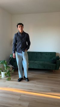 Svart skjorte med lange ermer, lys blå jeans og svarte sko. Står i et rom med en grønn fløyelssofa og planter.