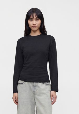 Ung kvinde med langt sort hår iført en ensfarvet sort langærmet trøje og lysegrå jeans mod en hvid baggrund.
