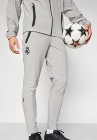 Hombre en pantalones de chándal grises del Real Madrid sosteniendo un balón de fútbol blanco con patrones de estrellas negras y rojas, usando botas negras y calcetas blancas.