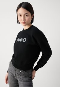 Zwarte gebreide trui met een zilveren "HUGO" logo op de voorkant. Gecombineerd met grijze denim jeans en een zwarte riem. Soepele textuur en slim fit ontwerp.