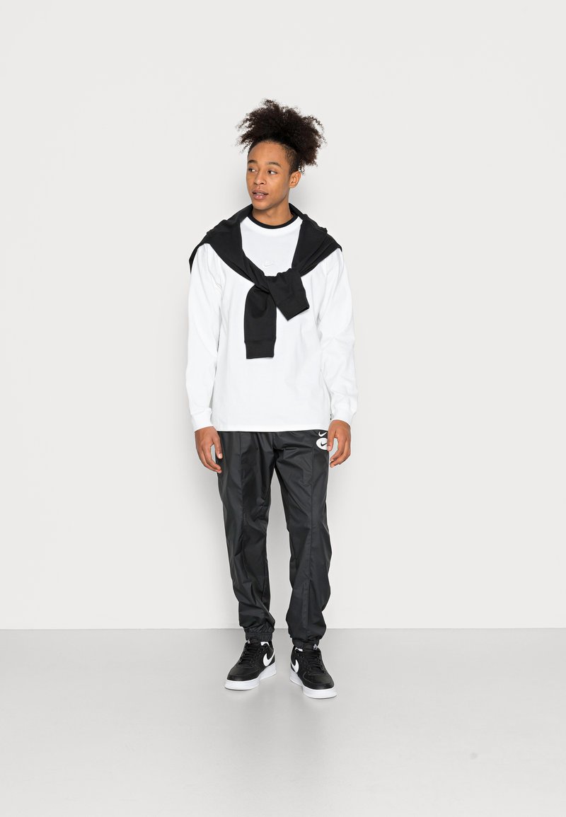 Witte lange mouw shirt gelaagd met een zwarte trui, gecombineerd met zwarte sportbroek met een logo, en zwarte sneakers met witte accenten.