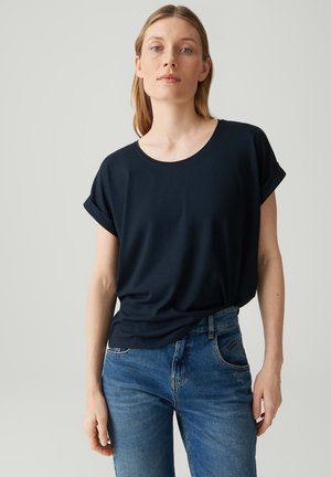 Jonge vrouw met licht haar draagt een effen donkerblauw T-shirt met opgerolde mouwen en blauwe spijkerbroek, staand tegen een effen achtergrond.