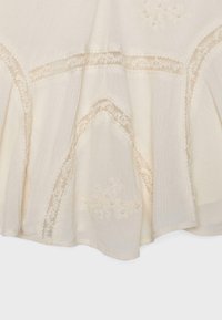 Tissu crème avec panneaux en dentelle transparente et broderie florale délicate formant des motifs courbes et linéaires.