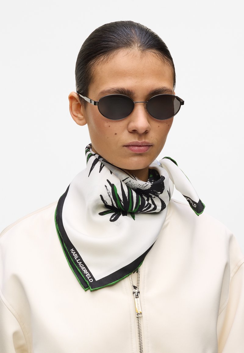 Γυναίκα με τα μαλλιά της προς τα πίσω, που φοράει μαύρα οβάλ γυαλιά ηλίου, λευκό μπουφάν με φερμουάρ και λευκό μεταξωτό μαντήλι με μοτίβα μαύρων και πράσινων φύλλων.
