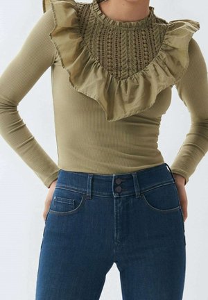 Kvinde iført olivengrøn ribbet top med lange ærmer, blonder og flæsedetaljer, kombineret med højtaljede mørkeblå jeans.