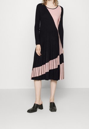 Vrouw die een zwart-roze geplooide midi-jurk met lange mouwen met diagonale kleurvlakken draagt en zwarte slip-on hakken, staand op een grijze vloer.