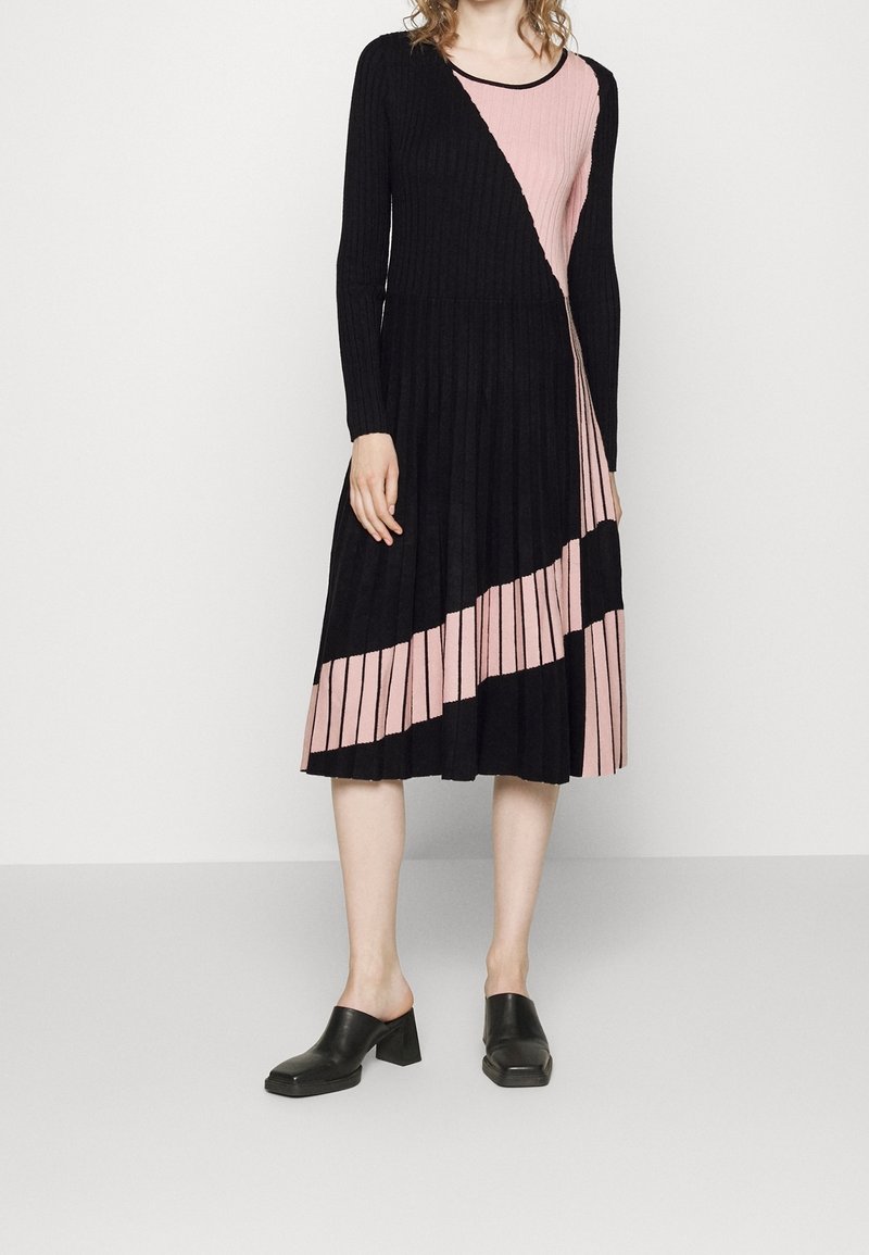 Femme portant une robe midi plissée à manches longues noire et rose avec des blocs de couleur diagonaux et des chaussures noires à talons plates, debout sur un sol gris.