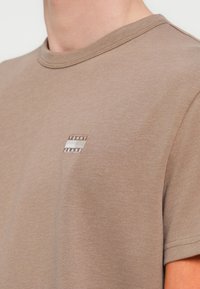 Καφέ βαμβακερό t-shirt με στρογγυλή λαιμόκοψη, με μικρό έμπλαστρο λογότυπου σε λευκό και μαύρο που γράφει "TOMMY JEANS" στο αριστερό στήθος.