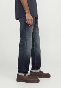 Tume sinine denim teksad kergelt kulunud efektiga ja rullitud äärega, koos pruunide nahkkingade ja tumesinise särgiga.