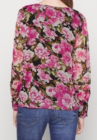 Blusa floreale con fiori rosa e bianchi su sfondo nero, tessuto trasparente, maniche lunghe e polsini con bottoni, abbinata a jeans blu.