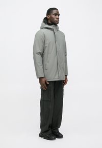 Sivá zateplená bunda s kapucňou, zapínaním na gombíky a bočnými vreckami, v kombinácii s čiernymi cargo nohavicami a tmavými topánkami. Jednoduchý, utilitárny dizajn.