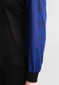 Giacca a maniche lunghe realizzata in tessuto perforato nero e con maniche blu con testo nero. I polsini a coste migliorano la vestibilità e la texture.