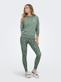 Jeune femme en sweat à capuche vert, leggings imprimé léopard vert et baskets blanches, debout les mains derrière le dos, regardant sur le côté.