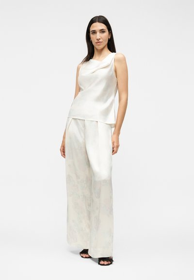 Γυναίκα με λευκό αμάνικο σατέν τοπ με ριγωτό ντεκολτέ και αντίστοιχη σατέν παντελόνα με φαρδιά πόδια, στέκεται μπροστά σε απλό φόντο.