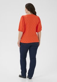 Orange texturerad topp med puffärmar och diamantmönster, paired med mörkblå jeans och svarta skor. Utsikt från baksidan.