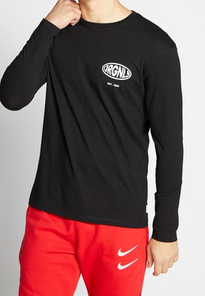 Osoba ubrana w czarną koszulę z długim rękawem z białym logo "ORGNLS EST. 1990" oraz czerwone spodnie z dwoma białymi logo Nike.