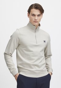 Light beige quarter-zip sweatshirt with ribbed collar and cuffs, featuring a small logo on the chest. Smooth texture and fitted design.