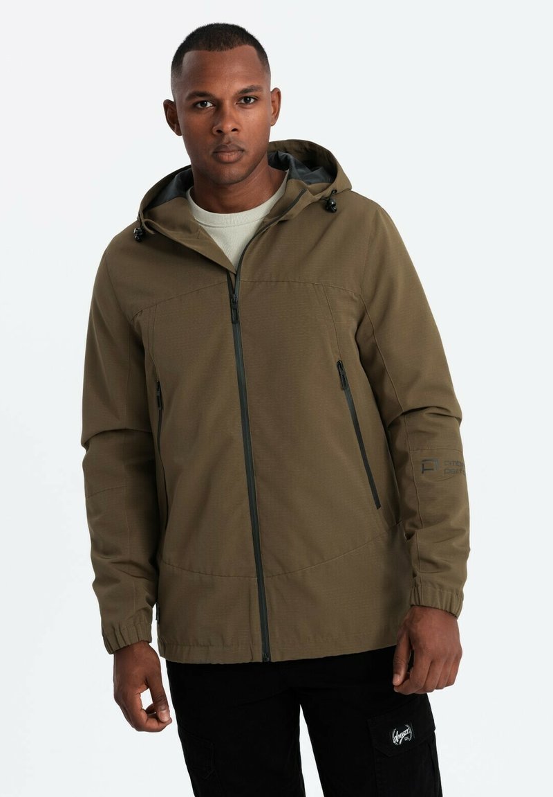 Veste à capuche vert foncé avec fermeture éclair à l'avant, poches latérales et poignets élastiques. Le tissu est lisse, avec une texture subtile et des lignes épurées.