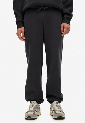 Person iført sorte joggerbukser med elastiske manchetter og lysegrå New Balance-sneakers, der står mod en hvid baggrund.