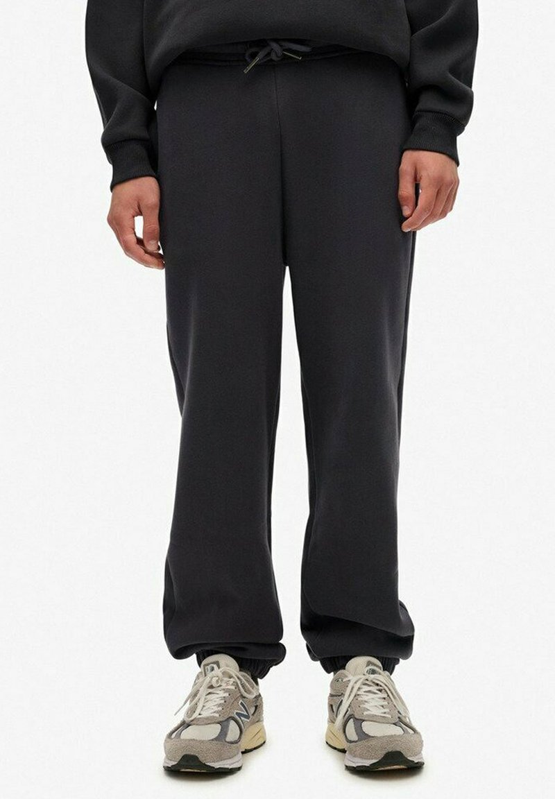 Personne portant un pantalon de jogging noir avec des poignets élastiques et des baskets New Balance gris clair, debout devant un fond blanc.