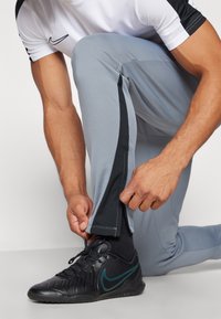 Grijze sportbroek met zwarte zijstrepen, gemaakt van lichtgewicht stof. Een persoon past het enkelgebied aan terwijl hij zwarte sneakers draagt.