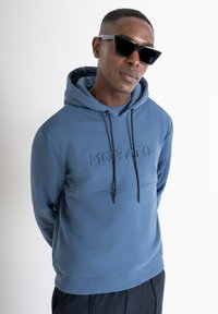 Felpa con cappuccio blu realizzata in materiale morbido con logo in rilievo, coulisse regolabile e polsini a coste; indossata con occhiali da sole scuri.