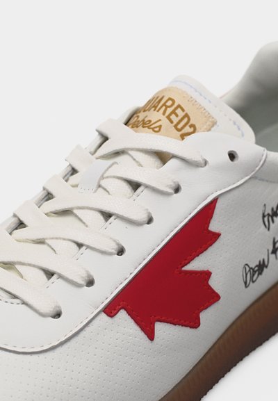 Λευκό αθλητικό παπούτσι με λευκά κορδόνια, καφέ σόλα, κόκκινη λεπτομέρεια με φύλλο σφενδάμου, ταμπά ετικέτα DSQUARED2 στη γλώσσα και μαύρο κείμενο με χαρακτηριστικό στυλ στην πλευρά.