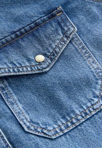 Denimskjortans fickdetalj, blått tyg, vit metalltryckknapp, kontrasterande orangea sömmar och synlig tygstruktur.