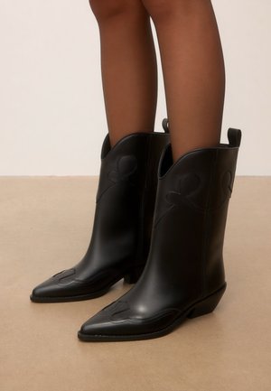 Vue en longueur de jambe d'une personne portant des bottes de cowboy en cuir noir mi-mollet avec des bouts pointus et des talons bas inclinés sur un sol beige.