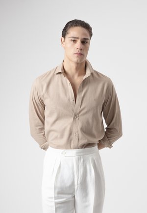 Hombre con el pelo peinado hacia atrás, vestido con una camisa beige abotonada y pantalones blancos de cintura alta, de pie con las manos detrás de la espalda.
