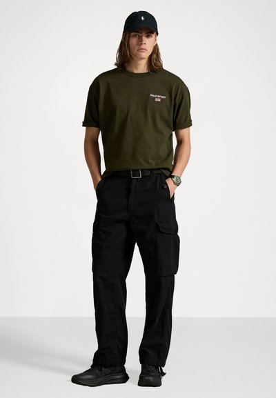 Olivegrünes Baumwoll-T-Shirt mit kurzen Ärmeln und kleinem Logo. Schwarze Cargo-Hosen mit Seitentaschen und verstellbaren Bündchen. Schwarze Sneakers und Mütze.