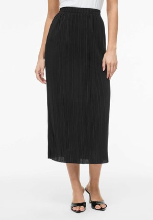 Femme portant une jupe midi plissée noire avec une ceinture élastique, associée à des sandales noires à talons ouvertes et un haut blanc.