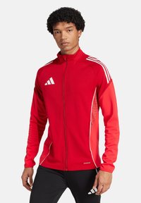 Rote Sportjacke mit durchgehendem Reißverschluss, aus glattem Stoff. Mit weißen Akzenten und drei weißen Streifen auf den Schultern.