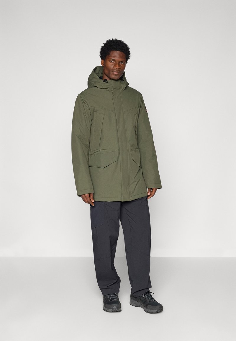 Parka kaki avec une capuche, dotée de grandes poches avant, d'une coupe droite et d'une texture lisse. Portée avec un pantalon sombre et des bottes.