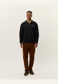 Sudadera negra con cremallera y cuello beige, combinada con pantalones de pana marrones y zapatos oscuros. El modelo está de pie frente a un fondo claro.