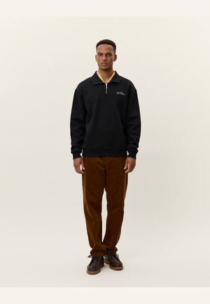 Sweatshirt noir zippé avec un col beige, associé à un pantalon en velours côtes marron et des chaussures foncées. Le mannequin se tient devant un fond clair.