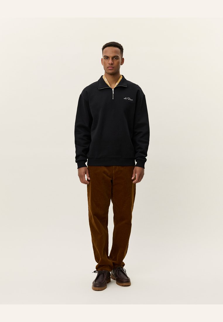 Felpa nera con zip e colletto beige, abbinata a pantaloni in velluto a coste marroni e scarpe scure. Il modello si trova di fronte a uno sfondo chiaro.