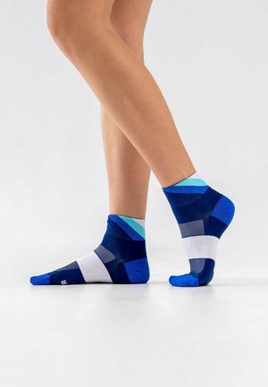Nackte Beine mit blau-weiß-türkis gestreiften Knöchelsocken, ein Fuß flach auf dem Boden, der andere auf den Zehenspitzen vor einem einfarbigen weißen Hintergrund.