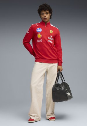 Nuori henkilö punainen Ferrari-brändätty kilpa-ajopaita päällä, leveät kermaiset housut, punaiset kengät, pitää mustaa Puma-duffel-laukkua harmaata taustaa vasten.