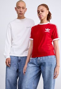 Crvena Adidas majica s vertikalnim prugama i bijelim detaljima, uparuena s laganim plavim trapericama. Model s lijeve strane nosi bijelu majicu s dugim rukavima.