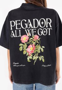 Juodas per didelis marškinėliai su gėlių grafika rožinėmis ir žaliosiomis spalvomis bei baltu užrašu: "PEGADOR ALL WE GOT" ir "Šeima & Draugai."