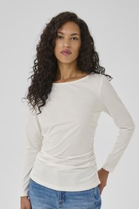 Woman with long curly hair wearing a white long-sleeve top and blue jeans, standing against a plain light gray background.