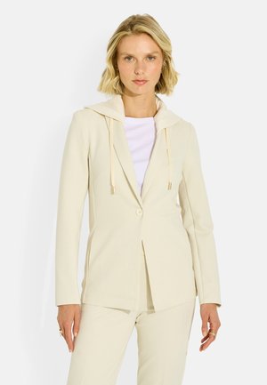 Mujer con un blazer color crema con capucha y pantalones a juego, de pie frente a un fondo blanco liso.