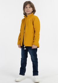 Gul zip-up jakke med stående krave, tætsiddende design og sidelommer. Båret sammen med mørkeblå jeans og hvide sneakers.