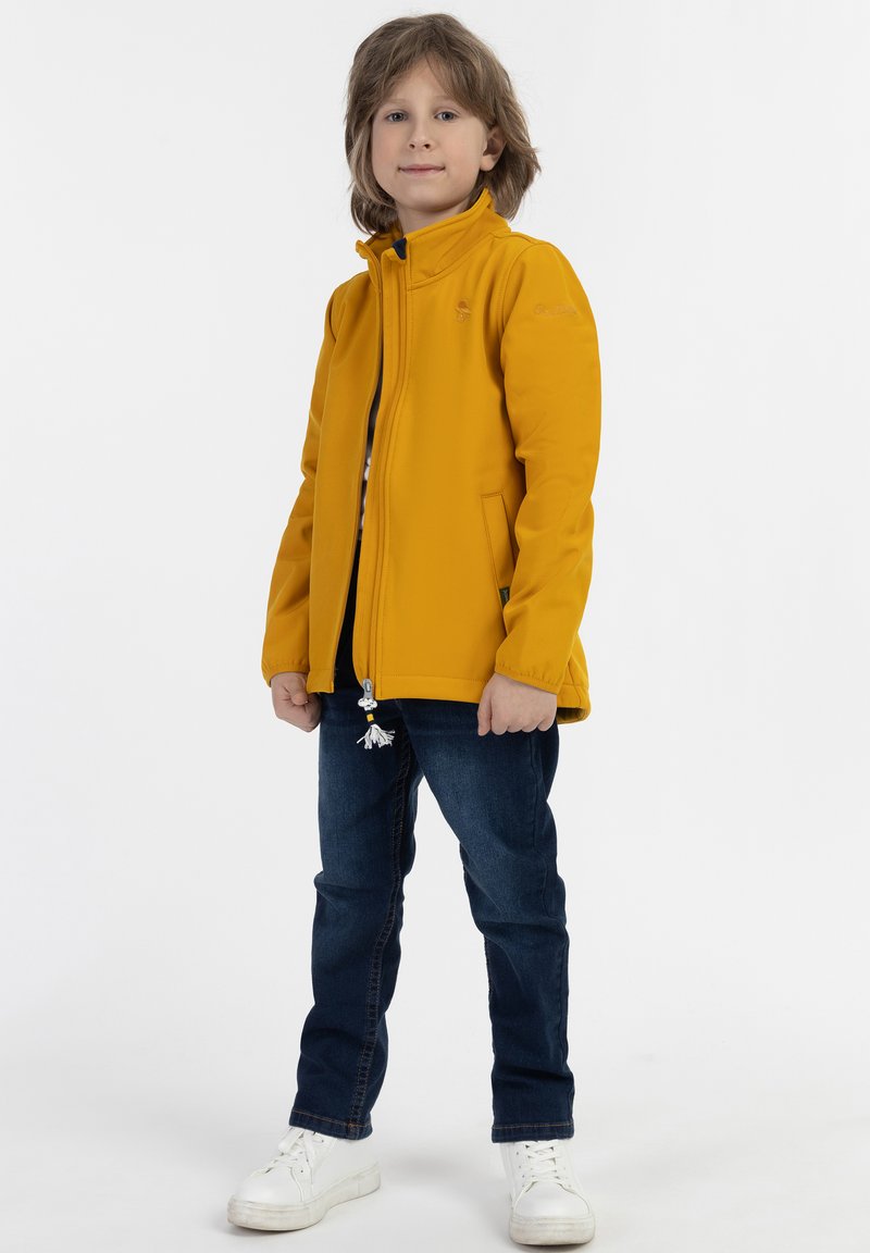 Gul zip-up jakke med stående krave, tætsiddende design og sidelommer. Båret sammen med mørkeblå jeans og hvide sneakers.