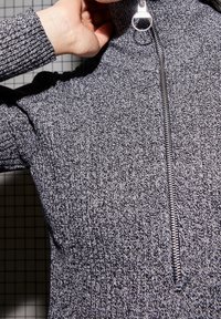 Suéter gris de punto con un alto cuello y media cremallera. Presenta un patrón texturizado y herrajes de cremallera plateados. Material suave y elástico.