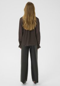 Pikkade blondide juustega naine, kes kannab läbipaistvat musta pluusi ja laiade jalgade musti nahkpükse, seisab valge tausta poole seljaga.